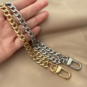 Puede incluir: Dos correas de cadena met&aacute;licas, una dorada y otra plateada, con cierres de mosquet&oacute;n. Las cadenas tienen un dise&ntilde;o de eslabones de bordillo grueso. Las correas se muestran sobre un fondo neutro.
