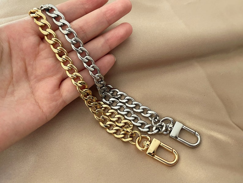 Puede incluir: Dos correas de cadena met&aacute;licas, una dorada y otra plateada, con cierres de mosquet&oacute;n. Las cadenas tienen un dise&ntilde;o de eslabones de bordillo grueso. Las correas se muestran sobre un fondo neutro.
