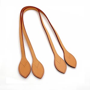 Puede incluir: Dos asas de bolso de cuero color canela con bordes marrón oscuro y costuras amarillas en contraste. Cada asa presenta dos pestañas de sujeción en forma de hoja en los extremos, perforadas con un patrón en V de pequeños agujeros.
