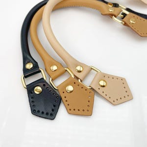 Könnte beinhalten: Vier Leder-Handtaschengriffe in Schwarz, Beige, Hellbraun und Beige. Jeder Griff hat ein geschwungenes Design mit goldfarbener Hardware und einer rautenförmigen Basis mit Stichlöchern. Die Griffe befinden sich auf weißem Hintergrund.