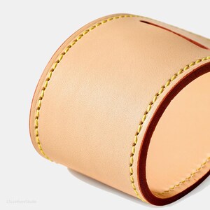 May include: Close-up of a handcrafted leather item, possibly a case or container. The item is made of light tan leather with a dark brown edge and yellow stitching. The design is simple and elegant, showcasing the quality of the materials.