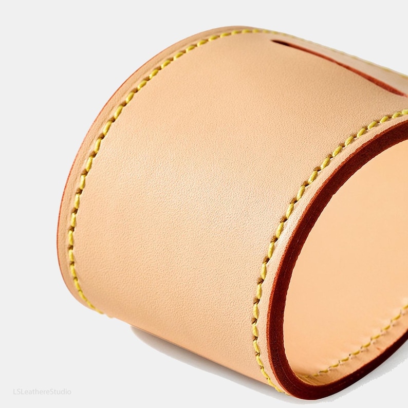 May include: Close-up of a handcrafted leather item, possibly a case or container. The item is made of light tan leather with a dark brown edge and yellow stitching. The design is simple and elegant, showcasing the quality of the materials.