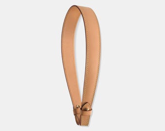 Op de afbeelding: Een natuurlijke Vachetta leren schouderriem. De riem heeft een lichtbruine kleur met een gladde textuur en zichtbare stiksels langs de randen. De riem heeft goudkleurige metalen hardware aan de onderkant. De tekst "Natural Vachetta Leather Shoulder Strap" staat bovenaan.