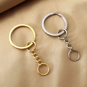 Könnte beinhalten: Zwei Schlüsselanhänger, einer in Gold und einer in Silber, mit einem runden Ring und einer Kette. Der goldene Schlüsselanhänger hat einen kleineren runden Ring am Ende der Kette.