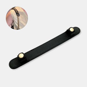 May include: Black leather shoulder strap protector with gold-colored button accents. The strap is designed to be placed on a shoulder to protect it from the weight of a chain strap. The protector is approximately 8 inches long.