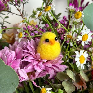 May include: A vibrant bouquet of flowers featuring a bright yellow felt chick perched on a pink chrysanthemum. The arrangement includes white daisies, purple flowers, and green foliage, creating a cheerful and colorful display.