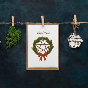 May include: A festive arrangement of holiday decorations hanging from a string. Includes a gift box, a sprig of greenery, a card with a pentagram wreath and the text "Blessed Yule!", a house-shaped ornament, and another gift box. All are attached with wooden clothespins.
