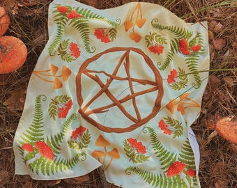 May include: A white cloth with a pentagram design in the center. The pentagram is made of brown branches. The cloth is decorated with red berries, green ferns, and brown mushrooms.