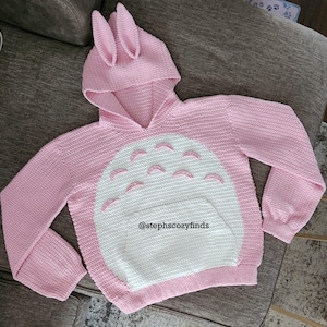 May include: A pink, handmade crochet hoodie with a bunny ear hood and a white belly design. The hoodie features a kangaroo pocket and is adorned with a design reminiscent of a popular animated character. The text "@stephscozyfinds" is visible on the front.
