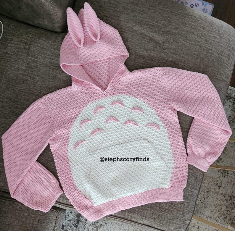 May include: A pink, handmade crochet hoodie with a bunny ear hood and a white belly design. The hoodie features a kangaroo pocket and is adorned with a design reminiscent of a popular animated character. The text "@stephscozyfinds" is visible on the front.