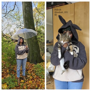 May include: A gray hooded sweatshirt with a Totoro design, featuring a cream-colored belly and a pocket. The hood has large, pointed ears. The person is wearing blue jeans and brown boots, holding a clear umbrella in a forest setting.