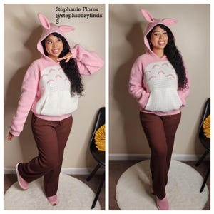 May include: A pink hooded sweater with bunny ears, a white pocket, and a cartoon design. The sweater is paired with brown pants and pink shoes. Text on the image reads "Stephanie Flores @stephscozyfinds S".