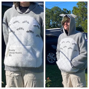 May include: A gray and white crochet hoodie with a character design. The hoodie features a large white front panel with a pocket and embroidered details. The hood has pointed ears. The text "Emily (they/them) XL" is visible.