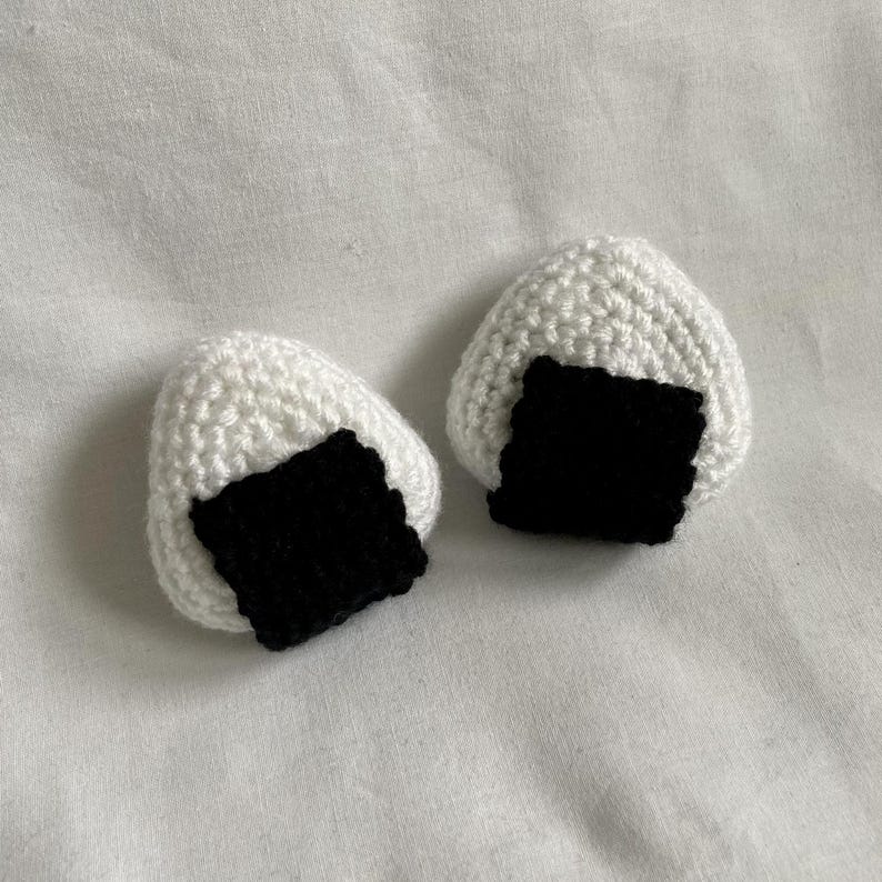 May include: Two white crocheted onigiri, a Japanese rice ball, with black crocheted nori seaweed.