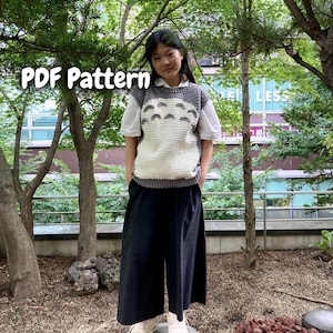 Op de afbeelding: Een persoon draagt een wit overhemd, een zwarte broek en witte sneakers, met een grijs en wit gehaakt vest. Het vest heeft een cartoonontwerp. De tekst "PDF Pattern" staat in de linkerbovenhoek.
