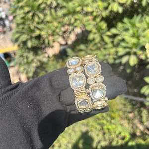 May include: Two gold bangles with large, clear, square-shaped gemstones set in the center of each link. The bangles are adorned with smaller, round, clear gemstones around the edges of the larger gemstones.