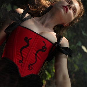 May include: A red corset with black straps and a snake design. The corset is worn by a person with long brown hair. The person is lying on their back with their head tilted back.