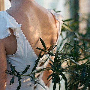 May include: A white dress with ruffled sleeves and a lace-up back is shown from behind. The dress is partially obscured by green foliage. The dress appears to be made of a soft, flowing fabric, and the overall aesthetic is romantic and ethereal.