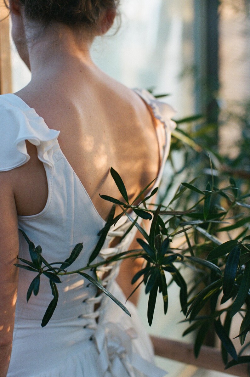 May include: A white dress with ruffled sleeves and a lace-up back is shown from behind. The dress is partially obscured by green foliage. The dress appears to be made of a soft, flowing fabric, and the overall aesthetic is romantic and ethereal.