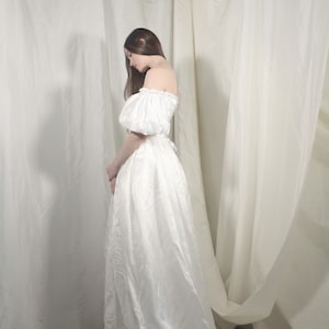 Puede incluir: Una mujer con un vestido blanco de hombros descubiertos y mangas abullonadas se encuentra frente a un fondo blanco.