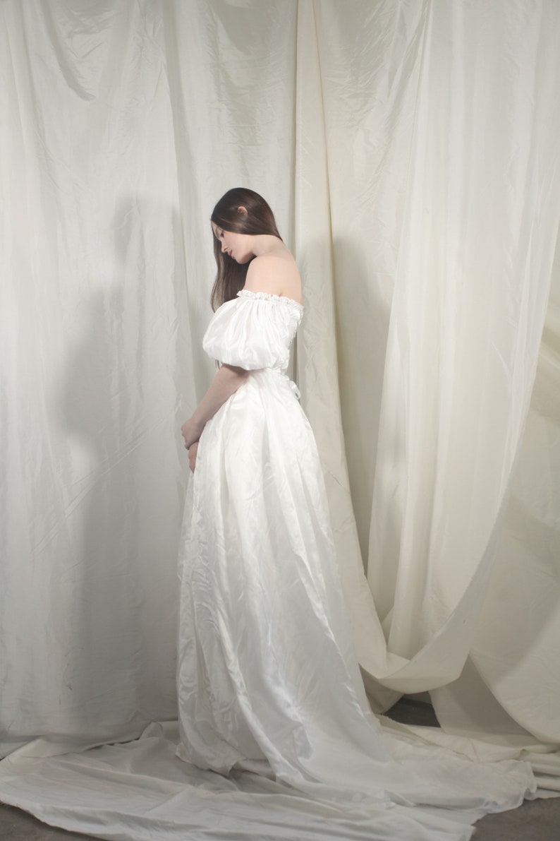 Puede incluir: Una mujer con un vestido blanco de hombros descubiertos y mangas abullonadas se encuentra frente a un fondo blanco.