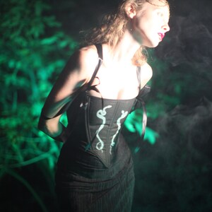 May include: A woman wearing a black corset with a white snake design. The corset has black straps and is worn over a black dress. The woman is standing in a dark, smoky environment.