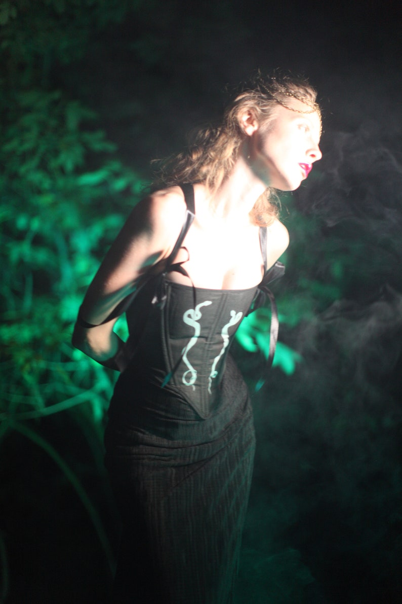 May include: A woman wearing a black corset with a white snake design. The corset has black straps and is worn over a black dress. The woman is standing in a dark, smoky environment.