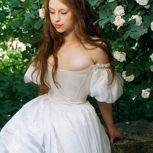 Puede incluir: Una mujer con un vestido blanco de hombros descubiertos con mangas abullonadas se encuentra en un jard&iacute;n con rosas blancas.