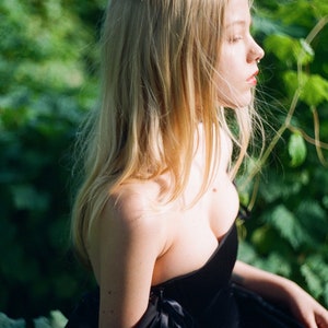 May include: A woman with long blonde hair wearing a black strapless dress with a sweetheart neckline. The dress has a black ribbon detail on the shoulder.