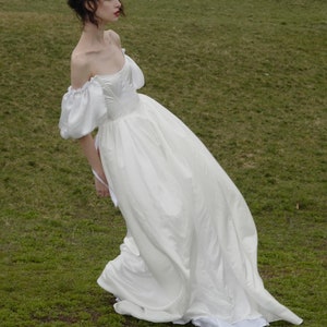 Puede incluir: Un vestido blanco de hombros descubiertos con mangas abullonadas y una falda fluida. El vestido est&aacute; fotografiado en una modelo de pie en un campo de hierba.
