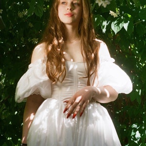 Puede incluir: Una mujer con un vestido blanco de mangas abullonadas se encuentra en un jard&iacute;n con hojas verdes y flores blancas. Tiene el pelo largo y casta&ntilde;o y lleva un anillo en el dedo.