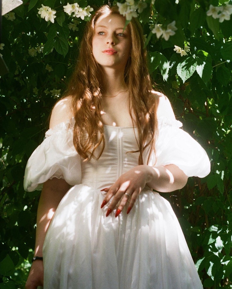 Puede incluir: Una mujer con un vestido blanco de mangas abullonadas se encuentra en un jard&iacute;n con hojas verdes y flores blancas. Tiene el pelo largo y casta&ntilde;o y lleva un anillo en el dedo.