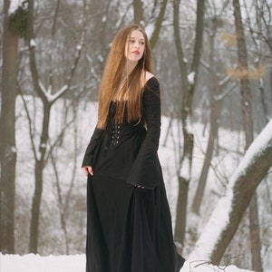 May include: A long-sleeved black floor-length dress featuring a square neckline, a fitted bodice with a central lace-up corset-style closure, and wide flared sleeves. The garment is constructed from a solid black fabric with a smooth texture.