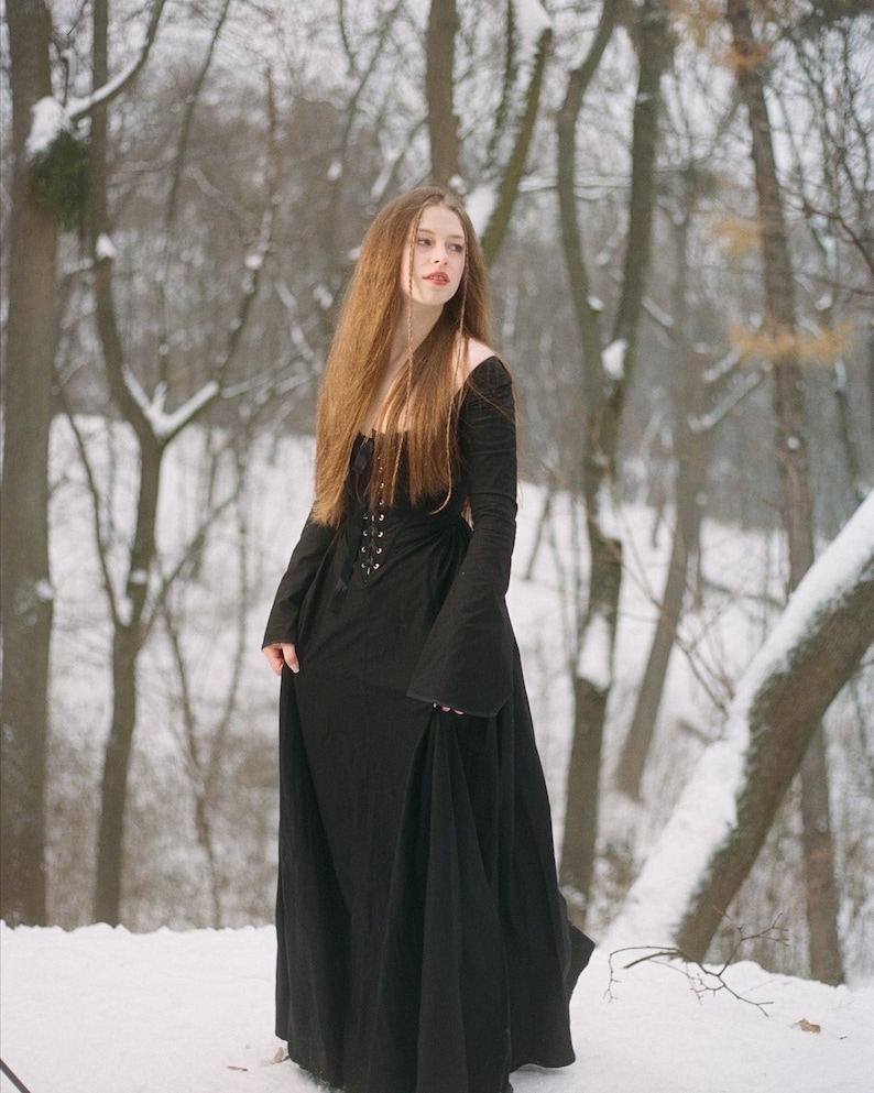 May include: A long-sleeved black floor-length dress featuring a square neckline, a fitted bodice with a central lace-up corset-style closure, and wide flared sleeves. The garment is constructed from a solid black fabric with a smooth texture.