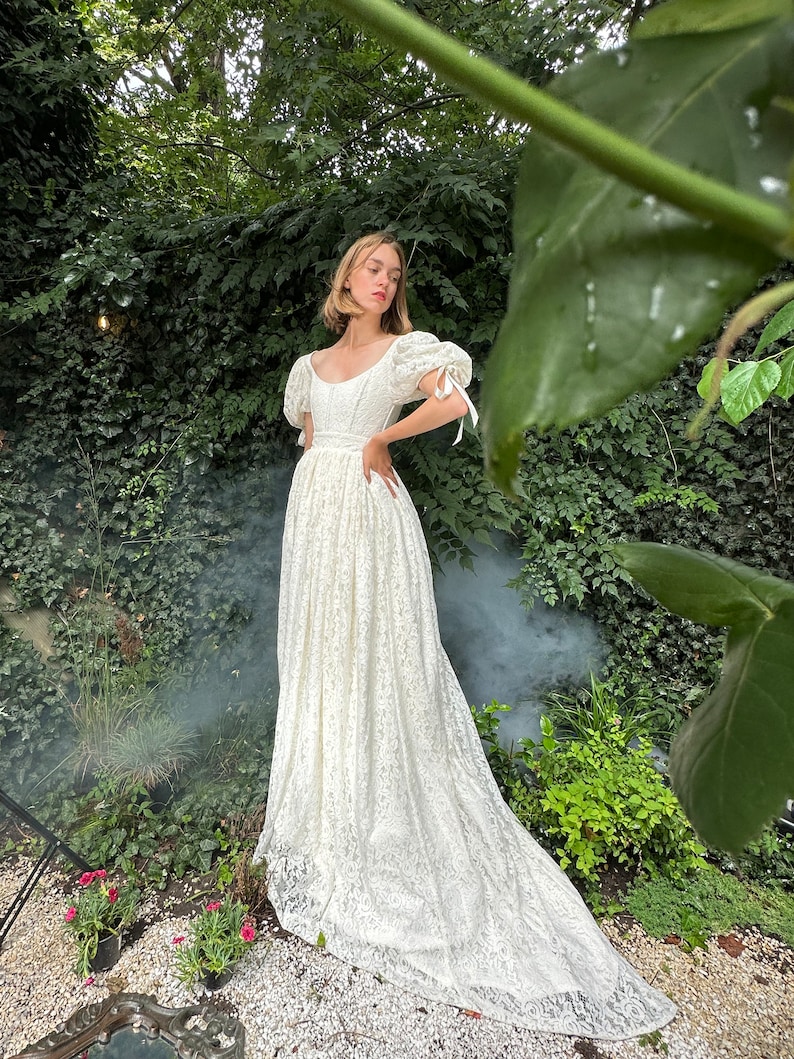 May include: A white lace wedding dress with a scoop neck, puff sleeves, and a long train. The dress is fitted at the bodice and flows out into a full skirt.