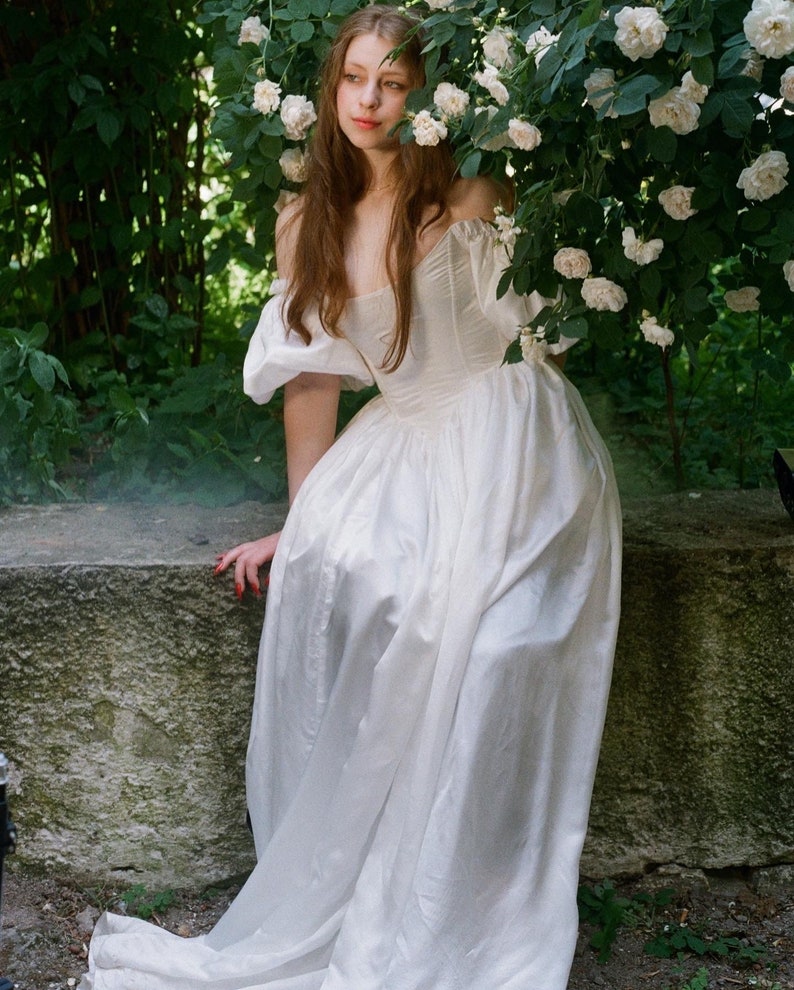 Puede incluir: Una mujer con un vestido blanco de hombros descubiertos con mangas abullonadas se sienta en un muro de piedra rodeado de rosas blancas.