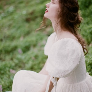 May include: A woman wearing a white lace dress with puff sleeves and a white ribbon tied at the wrist. The dress has a flowing skirt and a fitted bodice. The woman is sitting on a white surface with a green background.