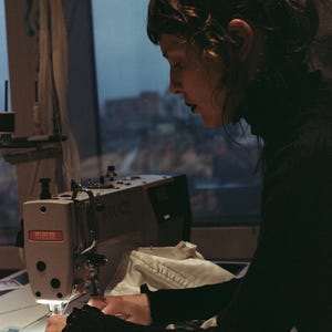 以下が含まれることがあります： 明るい光で照らされたミシンで作業する人。ミシンは薄いグレーで、「BRUCE」のブランド名が見えます。その人は黒い長袖のトップスを着ています。背景には窓から見える都市の風景があります。