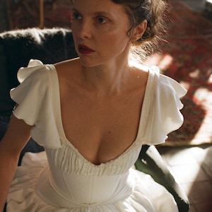 May include: A woman wearing a white dress with a low neckline and ruffled sleeves. The dress has a fitted bodice and a flowing skirt. The woman has dark hair and is wearing red lipstick. The background is blurred, with hints of a room.