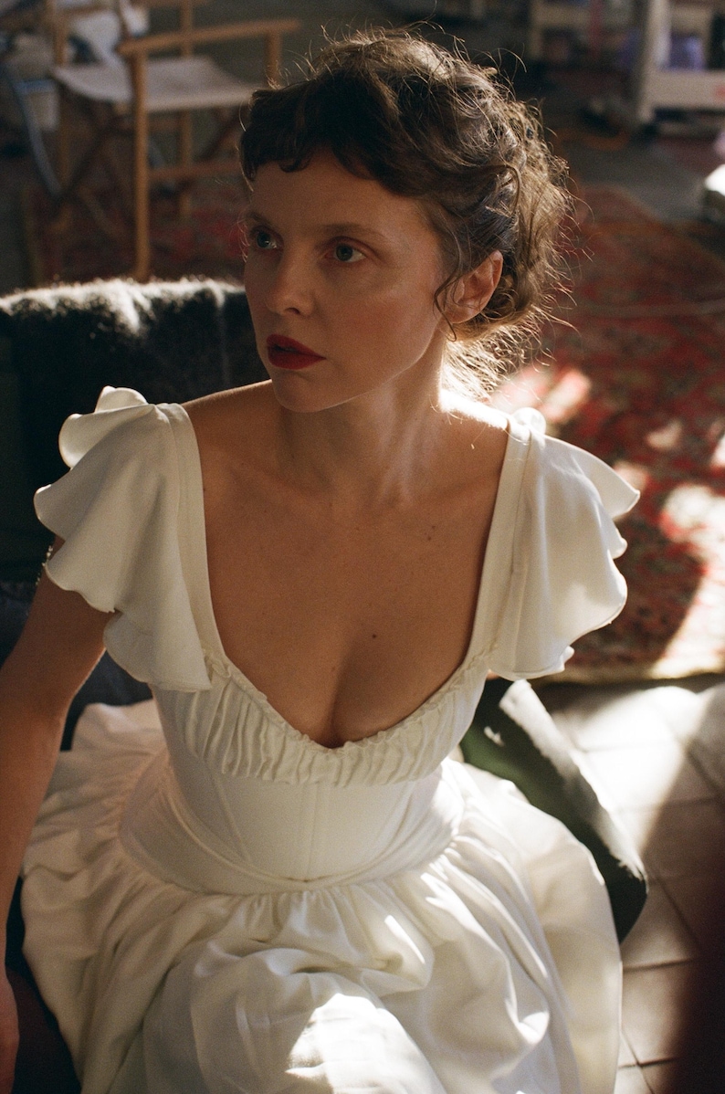 May include: A woman wearing a white dress with a low neckline and ruffled sleeves. The dress has a fitted bodice and a flowing skirt. The woman has dark hair and is wearing red lipstick. The background is blurred, with hints of a room.