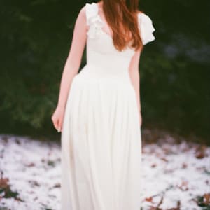 May include: A woman in a white dress stands in a snowy forest. The dress has a fitted bodice and a flowing skirt. The woman has long red hair and is looking down. The background is a blurred mix of green and white.