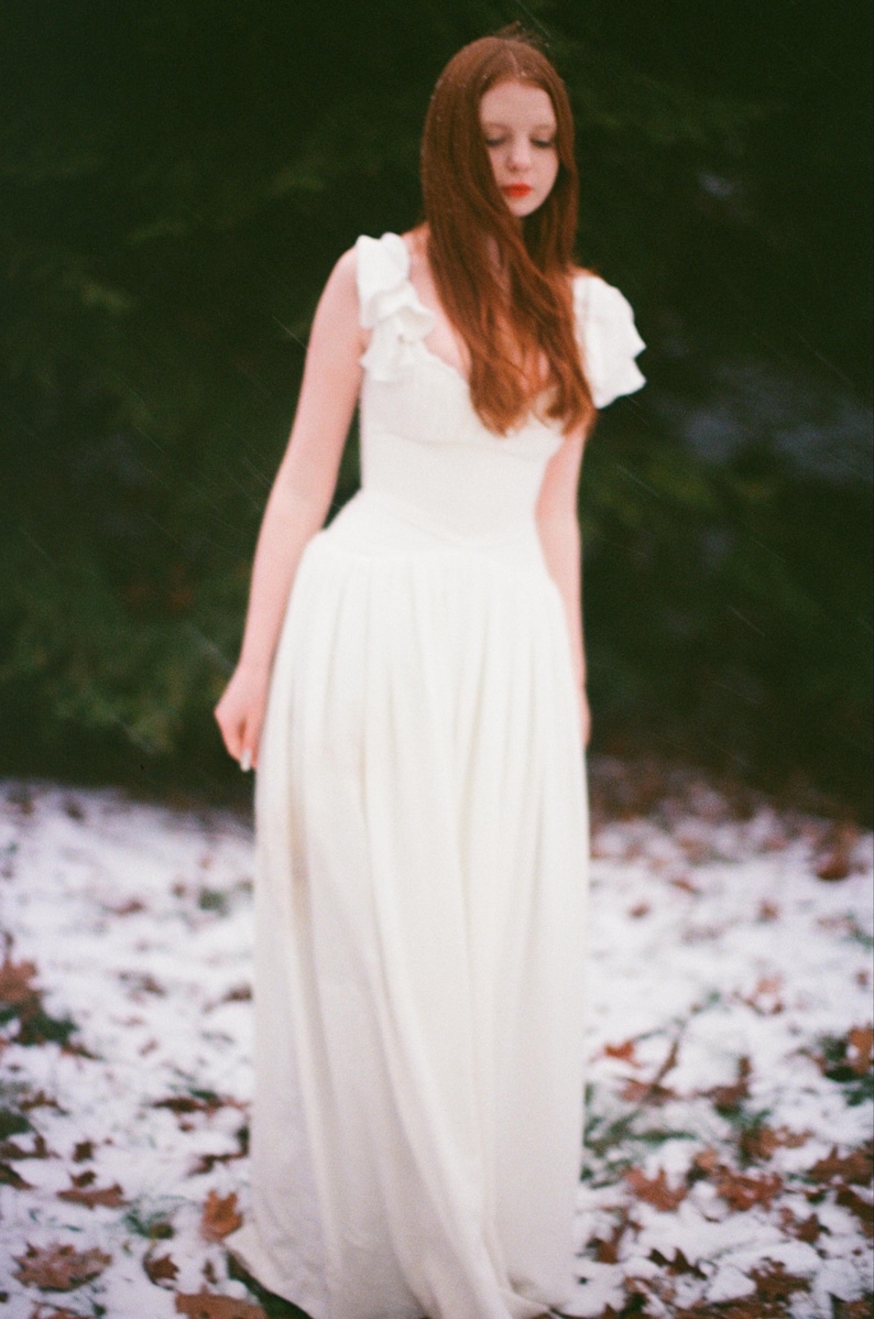 May include: A woman in a white dress stands in a snowy forest. The dress has a fitted bodice and a flowing skirt. The woman has long red hair and is looking down. The background is a blurred mix of green and white.