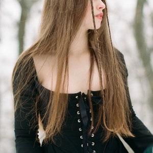 May include: A black long-sleeved dress featuring a square neckline and a front corset-style bodice with silver-toned metal eyelets and a black ribbon lace-up closure. The garment has a fitted torso and long sleeves, showcasing a solid black fabric.