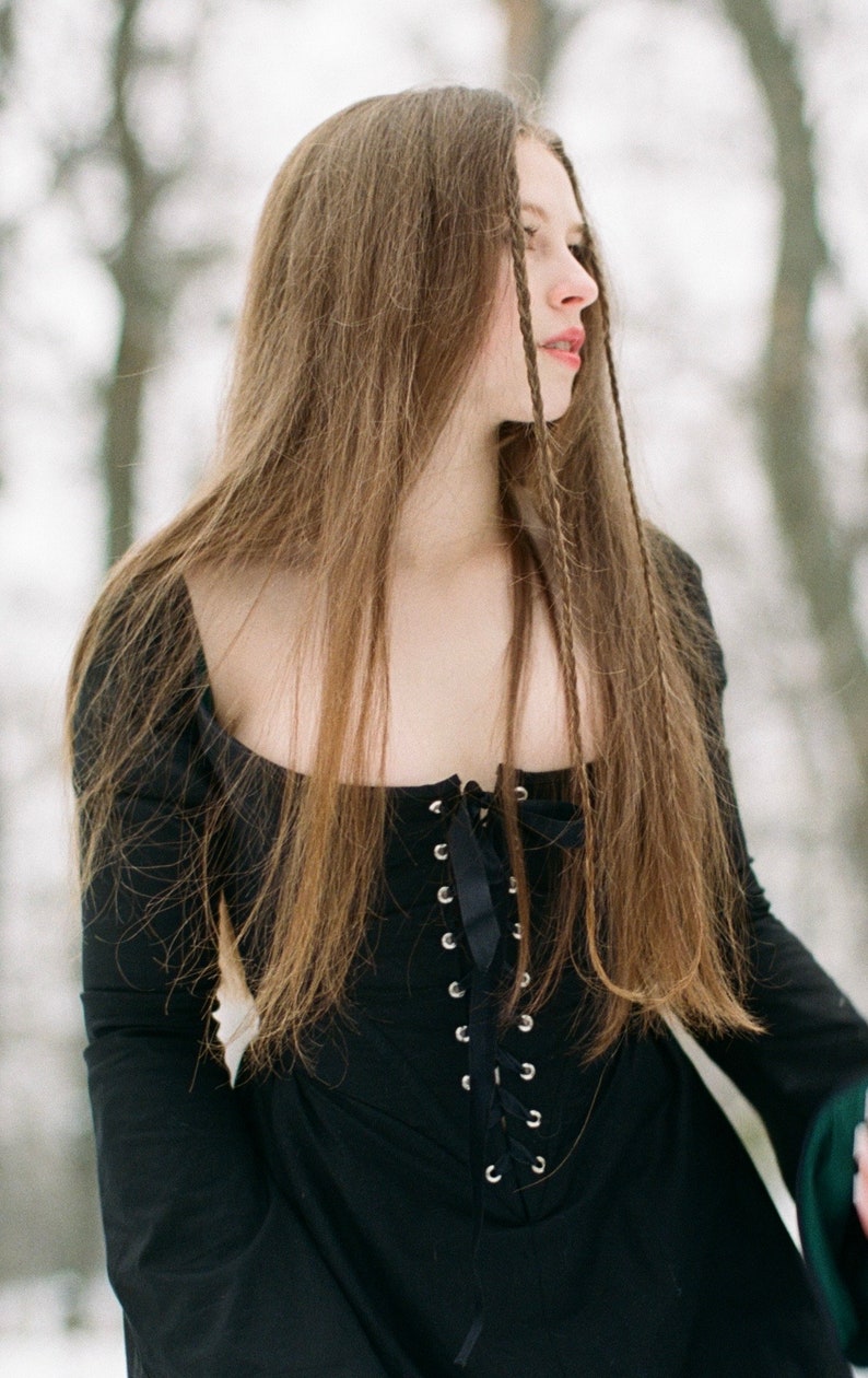 May include: A black long-sleeved dress featuring a square neckline and a front corset-style bodice with silver-toned metal eyelets and a black ribbon lace-up closure. The garment has a fitted torso and long sleeves, showcasing a solid black fabric.