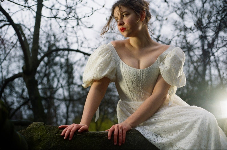 May include: A woman wearing a white lace dress with puffed sleeves. She is sitting on a tree branch with a forest background.