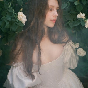Puede incluir: Una joven con el pelo largo y casta&ntilde;o lleva un vestido blanco de hombros descubiertos y se encuentra delante de un arbusto de rosas blancas.