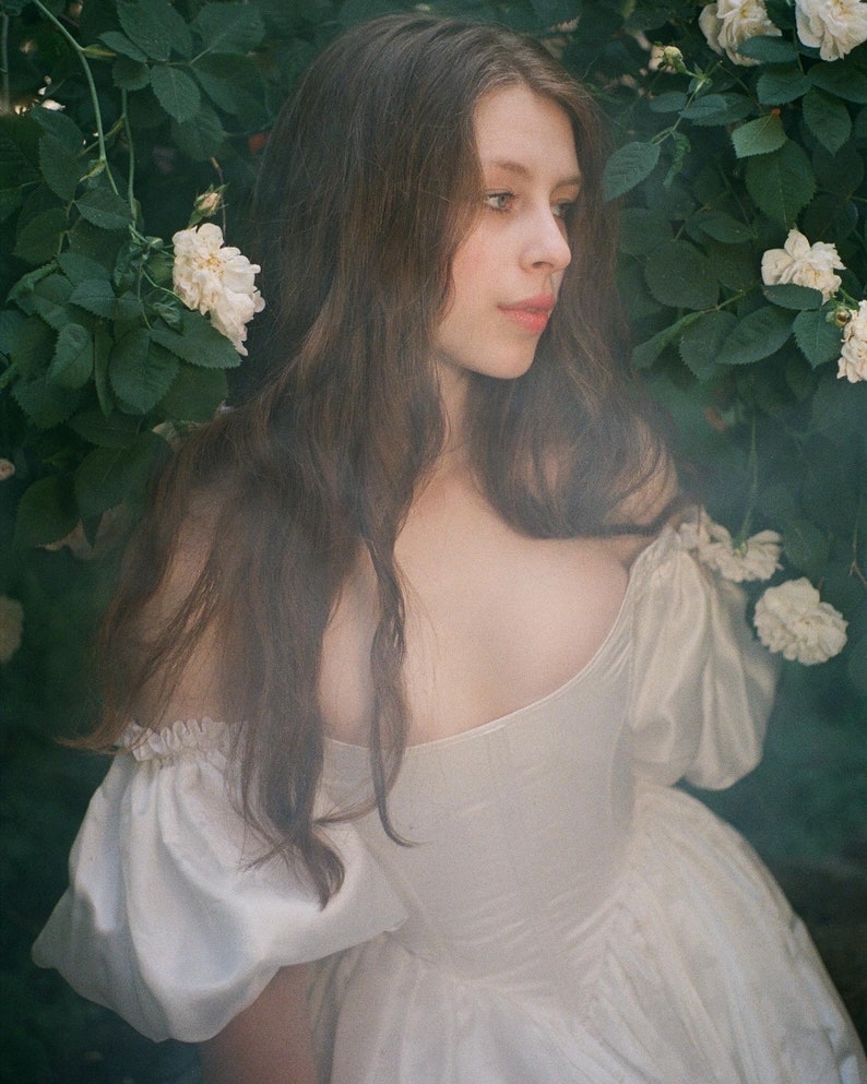 Puede incluir: Una joven con el pelo largo y casta&ntilde;o lleva un vestido blanco de hombros descubiertos y se encuentra delante de un arbusto de rosas blancas.