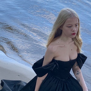 May include: A woman in a black off-the-shoulder gown with a fitted bodice and a flowing skirt. She is standing on a sandy beach with water in the background.
