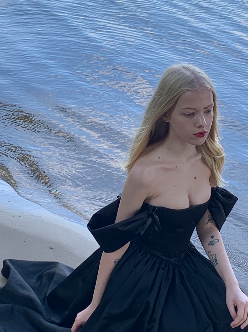 May include: A woman in a black off-the-shoulder gown with a fitted bodice and a flowing skirt. She is standing on a sandy beach with water in the background.
