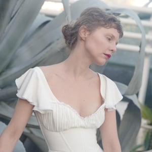 May include: A white dress with a fitted bodice and a flowing skirt. The dress features ruffled sleeves and a low neckline. The woman in the image has red lipstick and her hair is styled in a bun. The background includes large green plants.